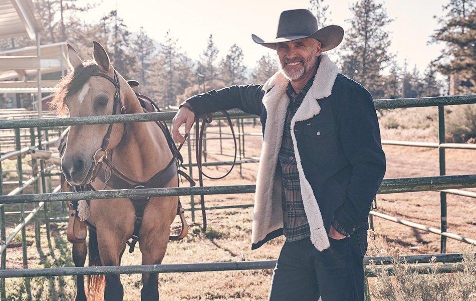 A man wearing sherpa jacket standing in a horse pen, smiling at the camera. Shop Sherpa