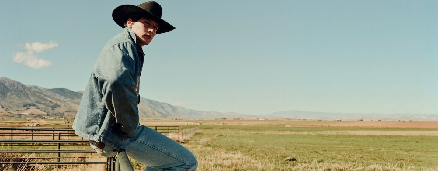 Closeup shot of a guy in a cowboy hat, sitting on a fence, looking off into a distant field.