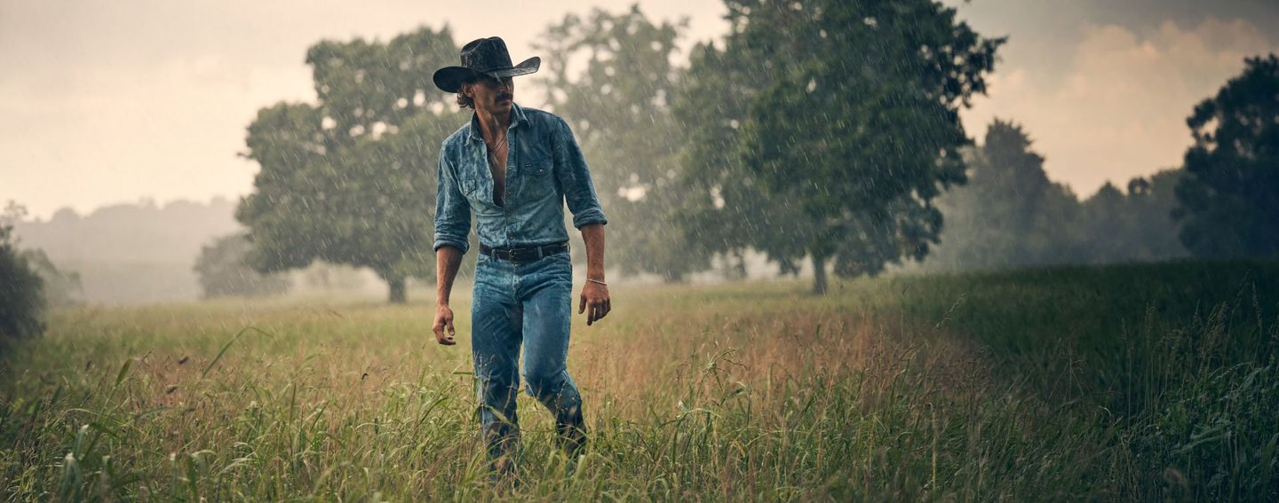 Tucker Wetmore walking through a field in the rain wearing a denim shirt and cowboy hat.