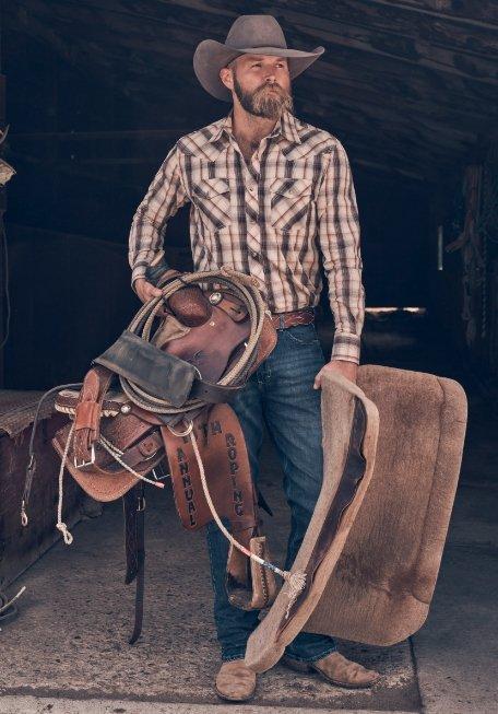 man holding a saddle and saddle pad wearing Wrangler apparel and a cowboy hat.