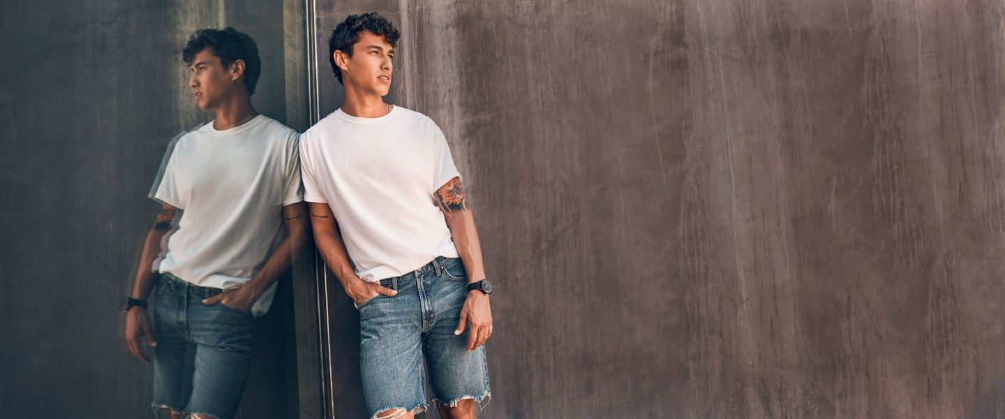 Guy in white tee shirt and denim shorts leaning against a concrete wall - shop shorts