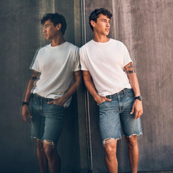 man in Wrangler denim shorts and white tee leaning up against window