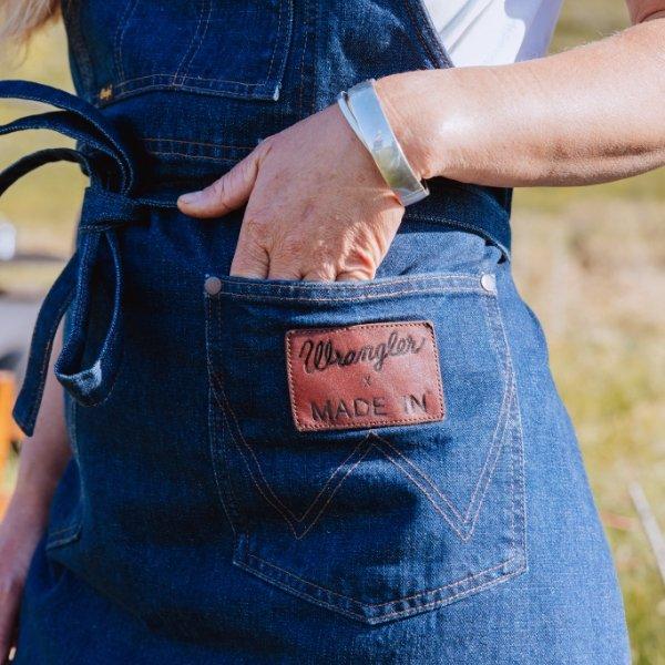 Person standing in a field wearing a denim apron that has a leather patch on it - Shop Wrangler x Made In
