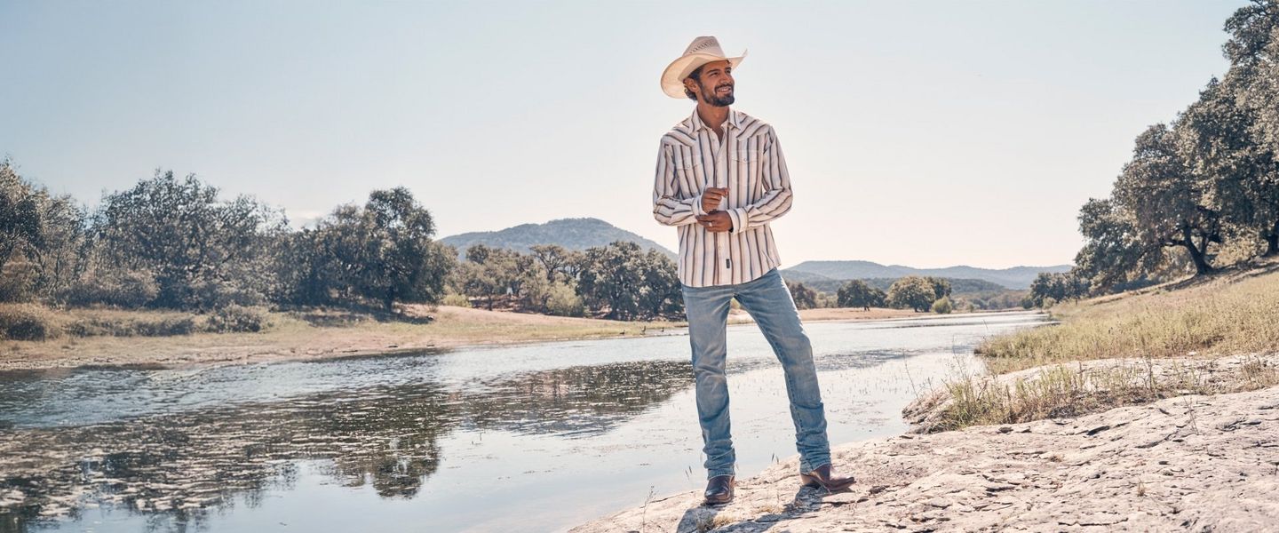 man wearing wrangler apparel and cowboy hat standing beside a river