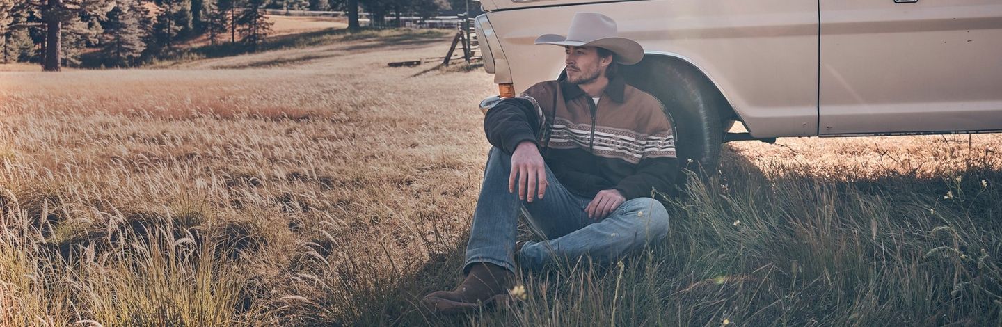 Guy sitting, leaned up against a truck in a field - Shop Jeans