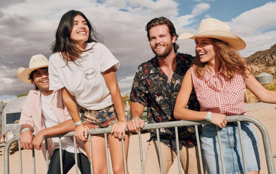 Four young adults smiling and leaning on a metal fence at an outdoor desert festival, wearing casual summer outfits and cowboy hats.
