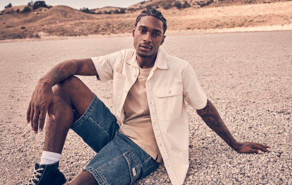 Man sitting on gravel in a desert landscape wearing a beige shirt and denim shorts.