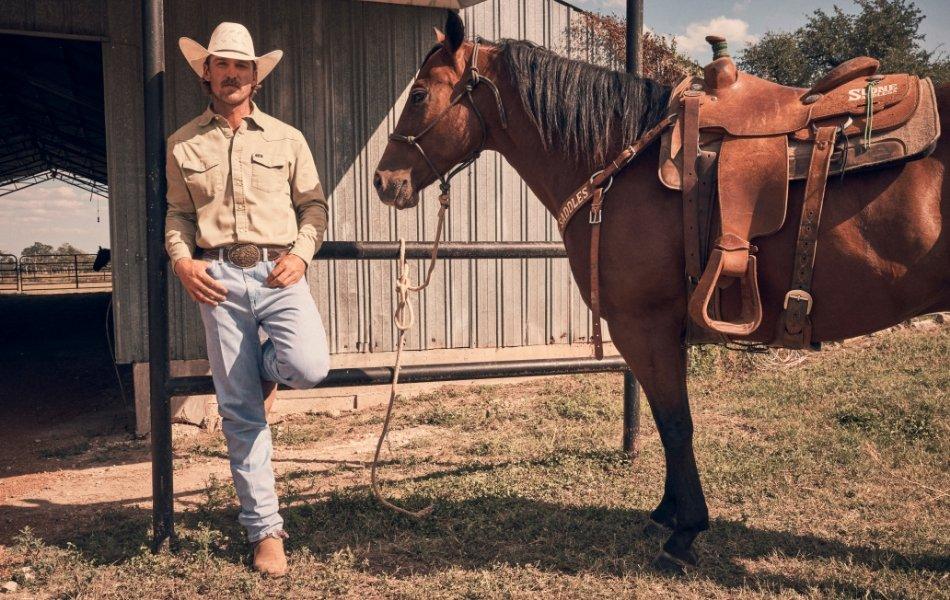 A man wearing Wrangler Cowboy Cut jeans and a cowboy hat standing next to his horse.