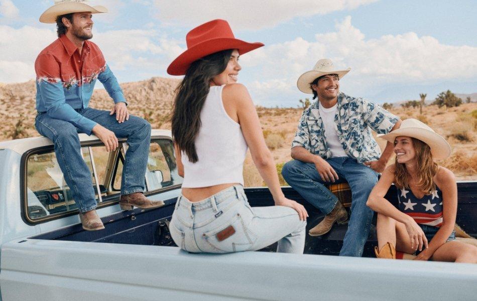 Four young adults smiling and sitting in a light blue truck bed, wearing casual summer outfits and cowboy hats.