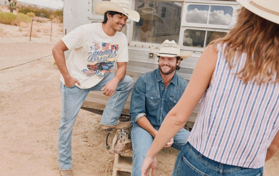 Three young men smiling and sitting beside a camper, wearing casual summer outfits and cowboy hats.