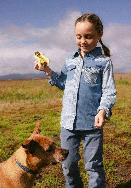 a girl wearing Wrangler apparel, holding a hotdog with a dog next to her