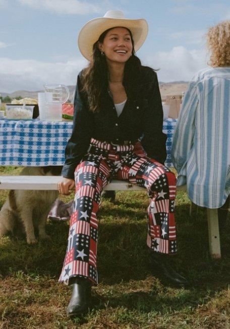 woman sitting on a bench wearing red, white and blue patterned pants and a cowboy hat