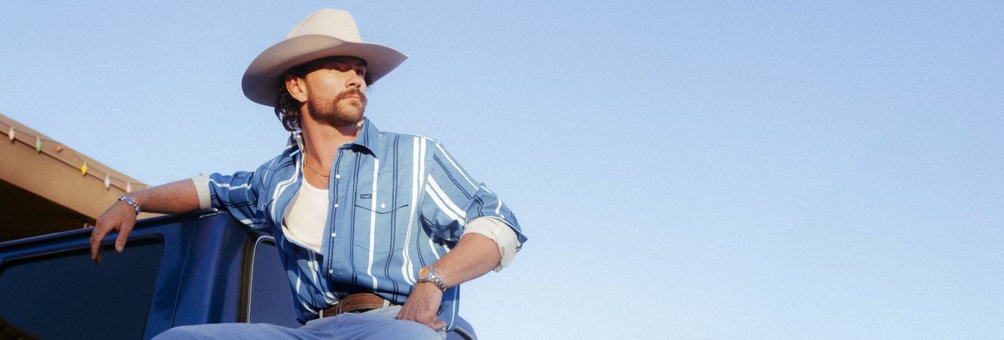 Guy in a cowboy hat sitting in the back of a truck with blue sky behind him - Shop Brushpopper