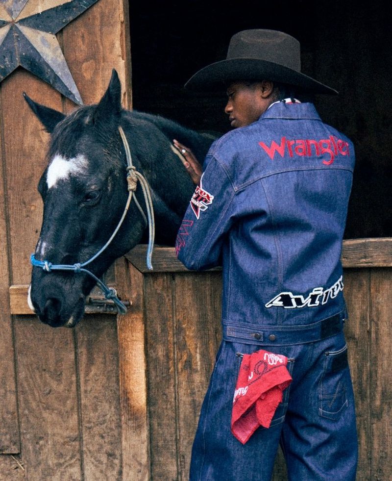 Man wearing Wrangler x Averix jacket and jeans, tending to a horse in a stall.